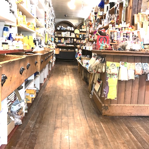 Looking down the long aisle of the store. Wide, worn oak floorboards run between cluttered shelves and built-in counters.