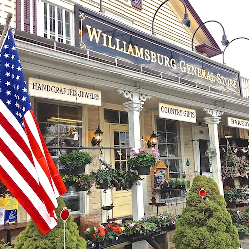 The Williamsburg General Store from Main Street. A long black sign reads WILLIAMSBURG GENERAL STORE in gold. American flag on the left. Hanging baskets of flowers along the porch. White-trimmed clapboard.
